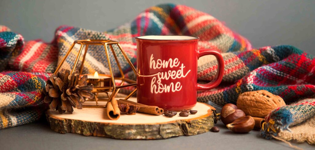 Picture of a red mug with home sweet home written on it beside a lit candle and throw blanket